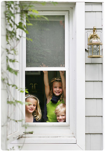 Three smiling children looking out of a window in the spring time.